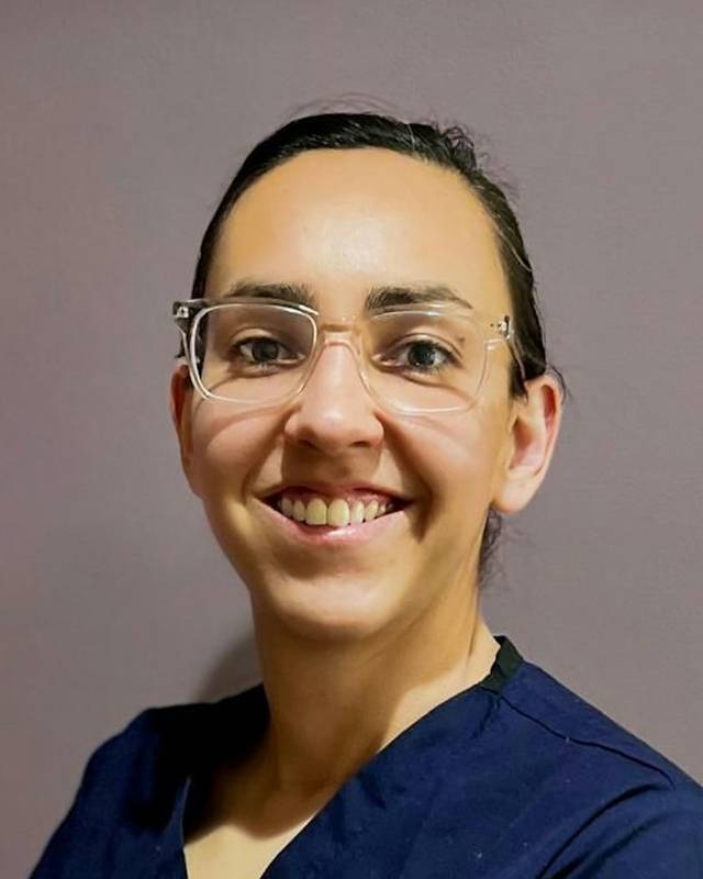 Retrato de la Dra. Clara Benlloch, mujer con gafas de montura transparente y pelo recogido, sonriendo con pijama sanitario azul marino.