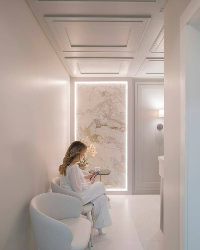 Sala de espera luminosa, con paredes claras y sillones blancos, donde una paciente lee mientras espera.